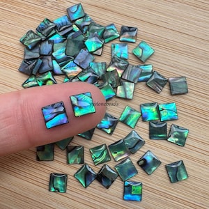 May include: A collection of iridescent green and blue abalone shell squares. The squares are approximately 1.2 cm in size and have a smooth, polished finish.