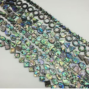 Może przedstawiać: Różnorodne, tęczowe koraliki z muszli abalone w różnych kształtach i rozmiarach, w tym kwadraty, owale, łzy i koła. Koraliki są nawleczone na białą sznurówkę i idealnie nadają się do tworzenia biżuterii.