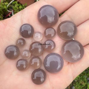 May include: A selection of polished, round, grey-purple gemstone beads in different sizes. The beads are smooth and reflective, with a translucent appearance. The image shows the beads held in a hand, with the text "yystonebeads" visible.