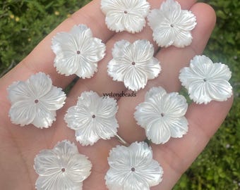 May include: A collection of eleven white, flower-shaped beads. Each bead has five petals and a small hole in the center. The beads have a pearlescent sheen, suggesting they are made of mother-of-pearl. These beads are ideal for jewelry making.