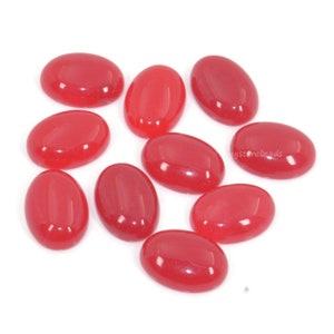 Puede incluir: Diez cabujones ovalados rojos, cada uno con un acabado liso y brillante. Los cabujones están dispuestos en un patrón disperso sobre un fondo blanco.