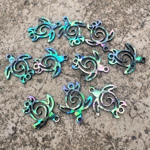 May include: A selection of iridescent sea turtle charms. Each charm has a stylized turtle design with a swirling shell pattern. The charms have a hole for stringing and display a range of colours, including blue, green, and purple.