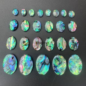 May include: An assortment of oval-shaped abalone shell cabochons in various sizes. The iridescent shells display a range of colors, including blues, greens, and purples, with a shimmering, textured surface. The cabochons are arranged on a dark background.