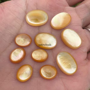 May include: A selection of oval and round, iridescent, golden-yellow cabochons. The cabochons have a pearlescent sheen and are of varying sizes. The text "yystonebeads" is visible on one of the cabochons.