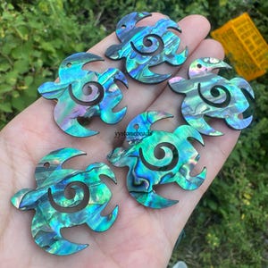 May include: A selection of iridescent abalone shell turtle pendants. Each pendant has a swirling design and a hole for threading. The shells show shades of blue, green, and purple. The text "yystonebeads" is visible.