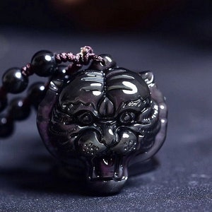Puede incluir: Colgante de cabeza de tigre de obsidiana oscura tallada. El colgante está ensartado en un collar de cuentas oscuras. Los rasgos del tigre están bien definidos, con ojos, nariz y boca visibles. El colgante mide aproximadamente 5 cm de largo.