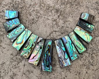 以下が含まれることがあります： 緑、青、金色のユニークなパターンが施された、虹色のアワビ貝のビーズ12個セット。ビーズは長方形で、滑らかで磨かれた仕上げになっています。黒いコードに糸を通しており、ジュエリー作りに最適です。