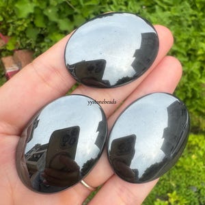 May include: Three polished, oval-shaped, black hematite stones. The stones are smooth and reflective, with a high-gloss finish. The stones are held in a hand, with the text "yystonebeads" visible.