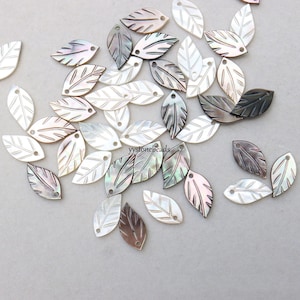 Puede incluir: Surtido de cuentas de concha en forma de hoja de color blanco, marrón e iridiscente con un agujero en la parte superior. Las cuentas están esparcidas sobre una superficie gris claro.