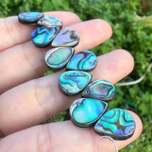 May include: A collection of iridescent abalone shell cabochons in shades of blue, green, and purple. The cabochons have a teardrop shape and are set on a dark base. The text "yystonebeads" is visible on one of the cabochons.