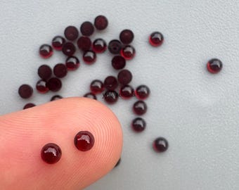 May include: A collection of small, round, deep red translucent beads scattered across a surface, with two beads resting on a fingertip. The beads have a smooth, polished finish and a dark garnet hue. The text ystonebeads is visible.
