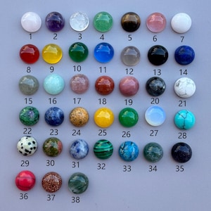 May include: A collection of thirty-eight round, polished cabochon gemstones arranged in a grid. Each stone features a unique colour, texture, or pattern, including solid, speckled, and marbled varieties, labelled with black numbers from 1 to 38.