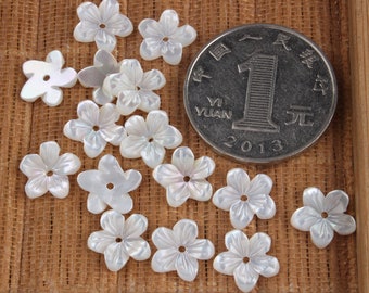 May include: A collection of white shell flower-shaped beads with a hole in the center. The beads are arranged on a woven surface with a Chinese yuan coin for scale.