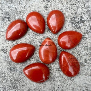 Könnte beinhalten: Acht rote Jaspis-Tropfencabochons, jeder mit einem einzigartigen Muster aus Streifen und einer glatten, polierten Oberfläche. Die Steine sind in einem Kreis auf einer grauen, strukturierten Oberfläche angeordnet.