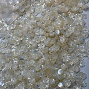 Puede incluir: Una gran cantidad de botones blancos iridiscentes en forma de flor. Los botones están hechos de nácar y tienen un diseño delicado y tallado.