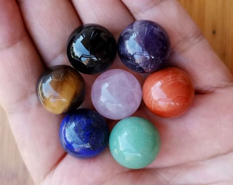 May include: A collection of seven polished gemstone spheres in various colors, including black, purple, brown, pink, orange, blue, and green. The spheres are held in a hand, showcasing their smooth, reflective surfaces. The gemstones are likely used for decorative or spiritual purposes.