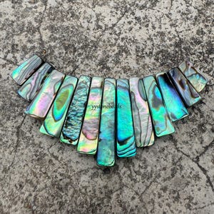 Könnte beinhalten: Eine Halskette aus irisierenden Abalone-Muschelteilen in Grün-, Blau- und Violetttönen. Die Teile sind fächerförmig angeordnet, wobei das größte Teil oben und das kleinste Teil unten ist. Die Halskette liegt auf einem grauen Hintergrund.