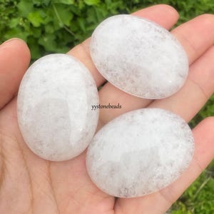 May include: Three smooth, oval-shaped white stones with subtle gray and pink veining are displayed. The stones are held in a hand, with the text "yystonebeads" visible. The stones appear to be polished and have a natural, organic aesthetic.