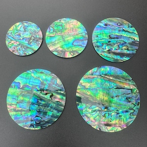 Könnte beinhalten: Fünf schillernde Abalone-Muschelscheiben in verschiedenen Größen. Die Muscheln zeigen eine lebendige Mischung aus Grün-, Blau- und Lilatönen mit natürlichen Mustern. Die Scheiben sind auf einer dunkelgrauen Oberfläche angeordnet.
