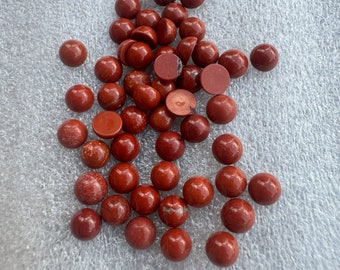 May include: A collection of polished, round, red jasper gemstones. The gemstones vary slightly in color and pattern, with some showing lighter veining. The stones are scattered on a textured, light gray surface.
