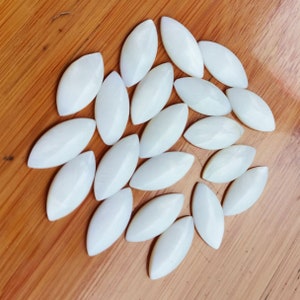 May include: A collection of 20 white oval-shaped mother of pearl shell cabochons. The cabochons are arranged in a loose circle on a wooden surface.