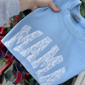 May include: A light blue sweatshirt with the word "MAMA" in large, white, floral-patterned letters. The sweatshirt is held by a person's hand, and the background includes red and blue floral decorations.