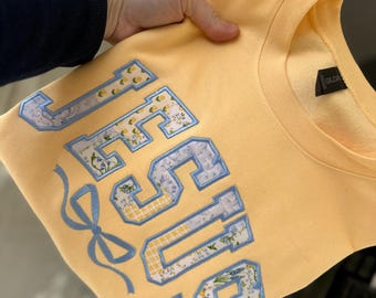 May include: A light yellow sweatshirt with the word "JESUS" in a collegiate style. The letters are outlined in blue and filled with floral and plaid patterns. A blue bow is embroidered below the letters.