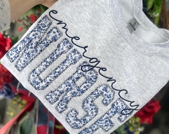 May include: Gray sweatshirt with the words "emergency nurse" embroidered in black with a blue floral pattern.