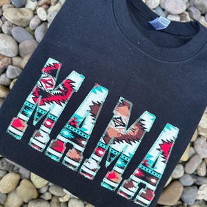 May include: Black sweatshirt with the word "MAMA" in large, white letters. The letters are decorated with a colorful, geometric pattern reminiscent of Native American designs.