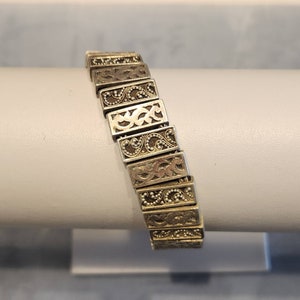 May include: A silver-toned stretch bracelet composed of rectangular metal links. Each link features an alternating pattern of embossed scrollwork and intricate beaded detailing, creating a textured surface across the entire length of the jewelry piece.