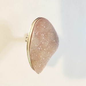 May include: A large, oval-shaped ring featuring a light pink, crystal-like stone with sparkling inclusions. The stone is set in a gold-colored metal band with a silver-colored adjustable ring base. The ring is displayed against a white background.