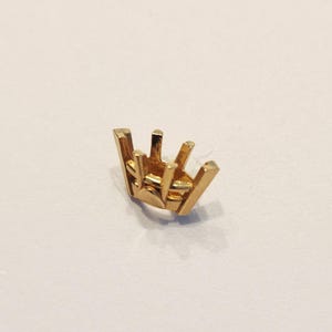 May include: A small, gold-colored jewelry setting with six prongs. The setting is designed to hold a gemstone. The prongs are evenly spaced and rise from a square base. The setting is on a white surface.