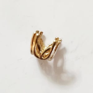 May include: A small, gold-colored jewelry finding with a floral design. The piece has a curved shape with multiple layers and is likely used in jewelry making or crafting. The finding is on a white surface.