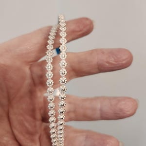 May include: A pair of thin, silver-toned bangles featuring a continuous, repeating floral or daisy-like pattern along the entire circumference of each band. The metallic surface has a bright, reflective finish with small, uniform circular details.