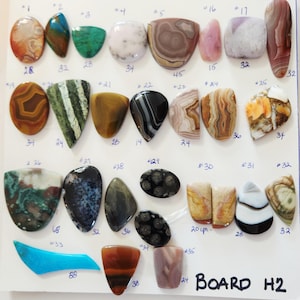 May include: A collection of polished gemstones in various shapes and colors, including oval, teardrop, and irregular forms. The gemstones are displayed on a white surface, with handwritten numbers and measurements in inches.
