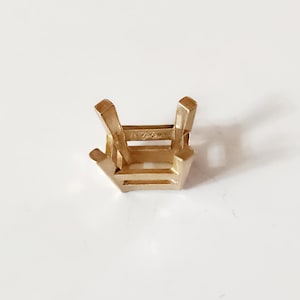 May include: A gold-colored jewelry setting with four prongs and a rectangular base. The setting is designed to hold a gemstone. The metal has a polished finish and is set against a white background.