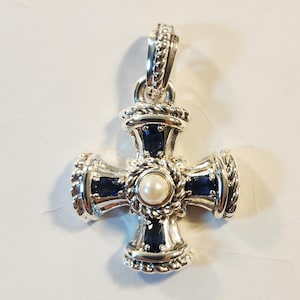 May include: A silver cross pendant featuring a central white pearl surrounded by a twisted rope design. Four oval-shaped dark blue sapphires are set into the arms of the cross, which also displays a textured rope border and a matching silver bail.