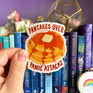 Könnte beinhalten: Ein weißer Aufkleber mit einer roten und gelben Illustration eines Stapels Pfannkuchen mit Butter und Sirup. Der Text "Pancakes Over Panic Attacks" ist in Rot auf dem Aufkleber gedruckt.