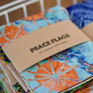 May include: A stack of colorful batik flags with a brown paper band that reads "PEACE FLAGS" and "10 unique batik flags". The flags feature orange and blue designs.