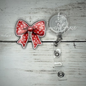 Puede incluir: Un lazo rojo y blanco con corazones blancos impresos en él está unido a un portagafete retráctil de plástico transparente. El portagafete tiene una correa de plástico transparente con un clip de metal y dos anillos de metal.