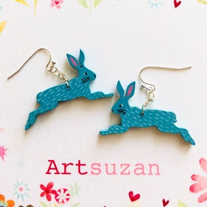May include: A pair of blue rabbit dangle earrings with pink accents. The earrings are handmade and feature a whimsical design.