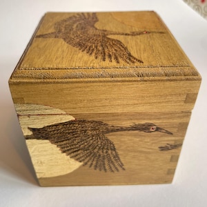 Puede incluir: Una caja de madera con tapa con bisagras, con un grabado detallado de una grulla en vuelo. La caja está hecha de madera de color claro, con el diseño de la grulla en un tono más oscuro. La caja tiene forma cuadrada.