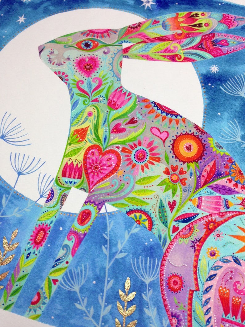 Op de afbeelding: Een kleurrijke, gedetailleerde illustratie van een konijn dat gesilhouetteerd is tegen een blauwe maan. Het konijn is bedekt met een patroon van bloemen, harten en paisley-ontwerpen in tinten roze, paars, groen en geel. De achtergrond is een lichtblauwe aquarelwas met gouden glitteraccenten.