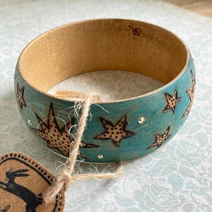 May include: A wooden bangle bracelet with a turquoise painted exterior. The bracelet features star designs with a brown, speckled pattern and small, clear rhinestones. A tag with a rabbit logo is attached with twine.