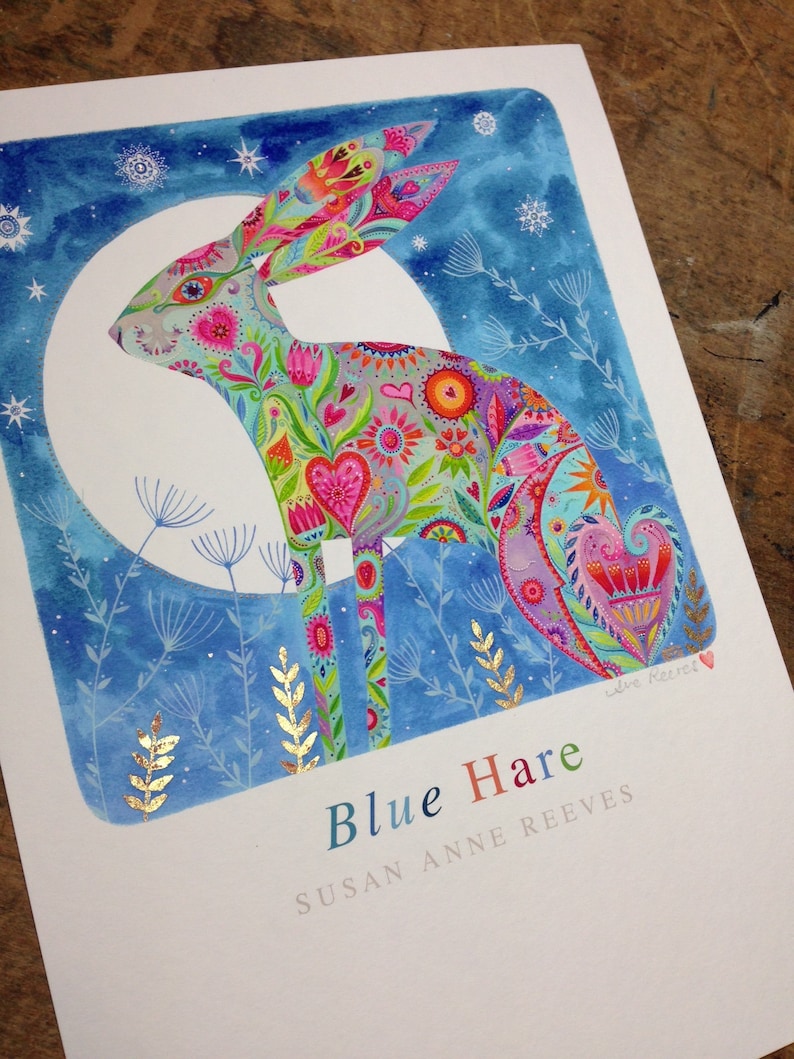 Op de afbeelding: Een kleurrijk aquarel schilderij van een blauwe haas met een maan en sterren op de achtergrond. De haas is gemaakt van verschillende bloemmotieven en harten. De tekst "Blue Hare" en "Susan Anne Reeves" staat onder de haas.