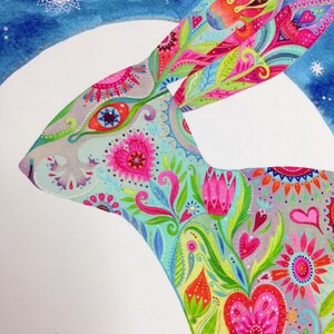 Op de afbeelding: Een kleurrijke, fantasierijke illustratie van een konijn dat gesilhouetteerd is tegen een witte maan. Het konijn is versierd met een levendig bloemenpatroon met harten, bloemen en paisley-ontwerpen in tinten roze, blauw, groen en geel. De achtergrond is een blauwe aquarelwas met witte sterren.