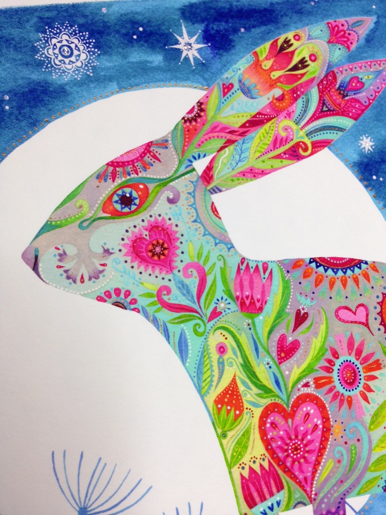 Op de afbeelding: Een kleurrijke, fantasierijke illustratie van een konijn dat gesilhouetteerd is tegen een witte maan. Het konijn is versierd met een levendig bloemenpatroon met harten, bloemen en paisley-ontwerpen in tinten roze, blauw, groen en geel. De achtergrond is een blauwe aquarelwas met witte sterren.