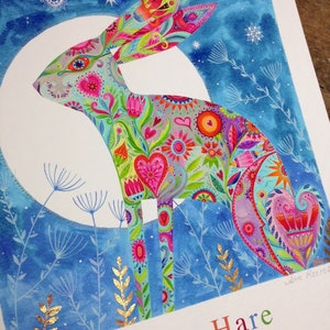 Op de afbeelding: Een aquarel schilderij van een blauwe haas met een kleurrijk bloemenpatroon. De haas staat op zijn achterpoten tegen een blauwe achtergrond met witte sterren. Het schilderij is gesigneerd met "Jane Reeves".