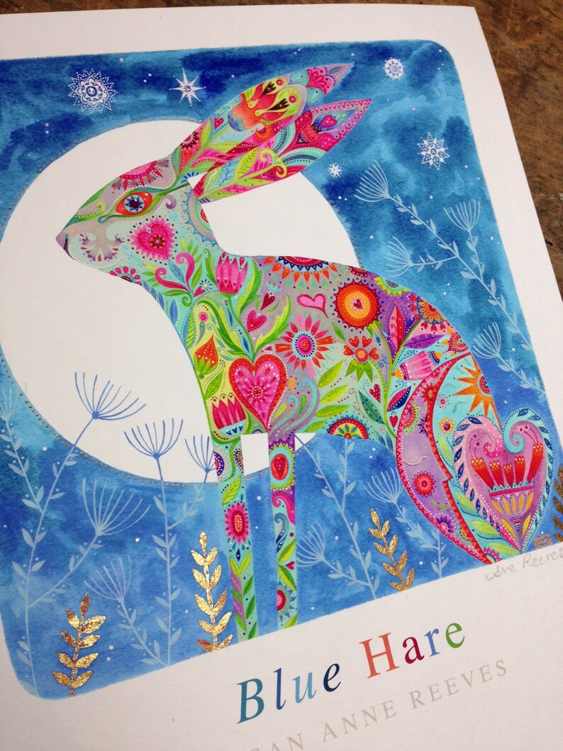 Op de afbeelding: Een aquarel schilderij van een blauwe haas met een kleurrijk bloemenpatroon. De haas staat op zijn achterpoten tegen een blauwe achtergrond met witte sterren. Het schilderij is gesigneerd met "Jane Reeves".