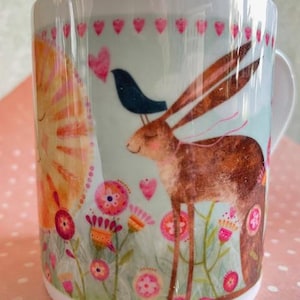 May include: White ceramic mug with a whimsical design. It features a brown hare with long ears, a blue bird, and colorful flowers. The mug has a light blue background with pink hearts and a sun-like figure. A decorative item.